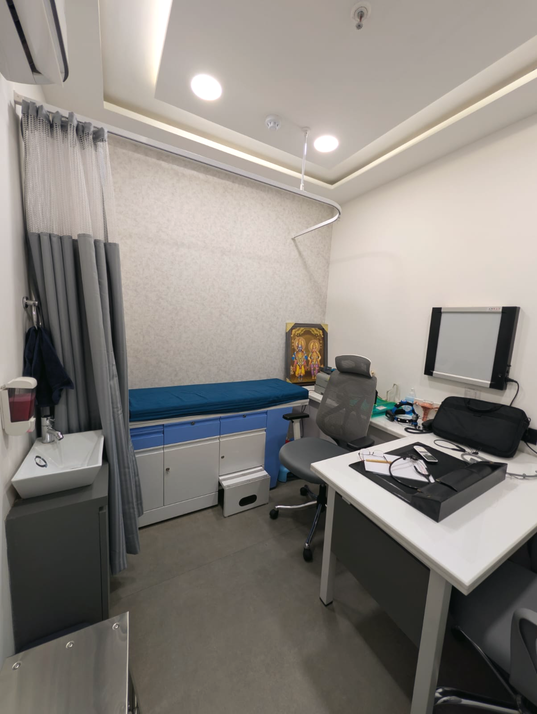 The consultation room at Doctors Planet NX, Hiranandani Estate — examination bed with curtain, desk, and warm overhead lighting.